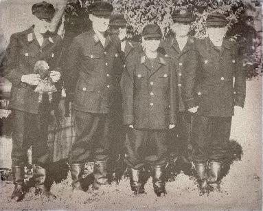 Sechs Personen in Uniform, mit Hüten und Stiefeln, stehen im Freien zusammen. Eine Person hält eine Puppe in der Hand. Das Schwarz-Weiß-Foto wirkt gealtert oder alt und bietet einen Einblick in die Geschichte der Wehr.