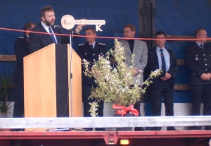 Ein Mann spricht an einem Podium auf der Bühne, hinter ihm stehen mehrere Personen. Davor steht ein kleiner Baum mit einem roten Band, und über dem Kopf hängt ein großer symbolischer Schlüssel - ein Ereignis, das Themen der Wehrgeschichte widerspiegelt.