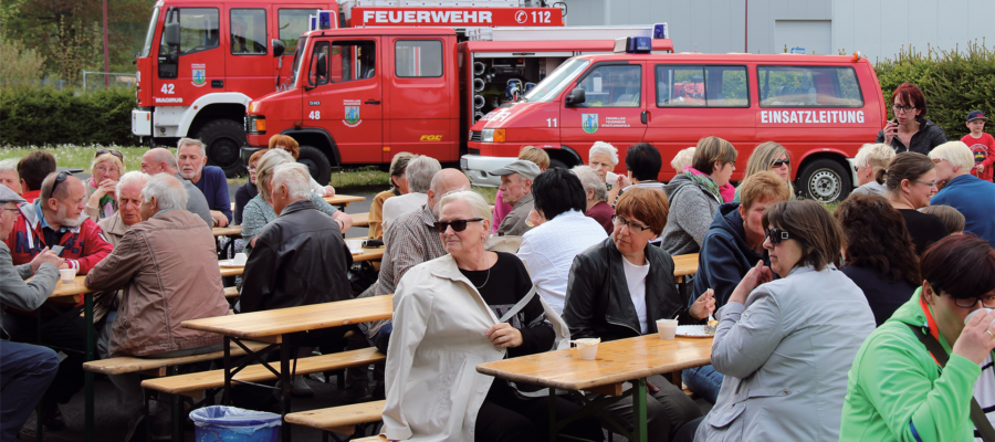 Eine Gruppe von Menschen sitzt an Picknicktischen im Freien, isst und unterhält sich. Im Hintergrund sind zwei rote Feuerwehrautos mit deutscher Aufschrift zu sehen, und ein paar Leute stehen in der Nähe der Fahrzeuge. Es scheint sich um eine Gemeinschaftsveranstaltung zu handeln.