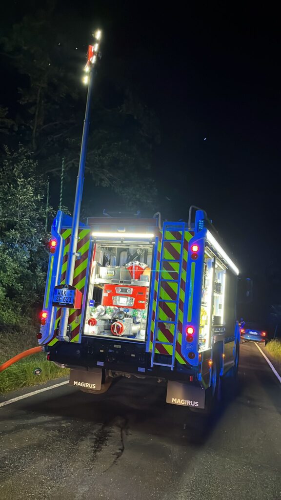 Ein hell erleuchtetes Feuerwehrauto ist nachts auf einer dunklen Straße geparkt. Die hinteren Abteile sind geöffnet und geben den Blick auf die Feuerwehrausrüstung frei. Ein großer Scheinwerfer ist ausgefahren und erhellt die Szene. Im Hintergrund sind Bäume zu sehen.