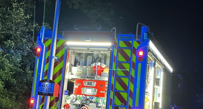 Ein hell erleuchtetes Feuerwehrauto ist nachts auf einer dunklen Straße geparkt. Die hinteren Abteile sind geöffnet und geben den Blick auf die Feuerwehrausrüstung frei. Ein großer Scheinwerfer ist ausgefahren und erhellt die Szene. Im Hintergrund sind Bäume zu sehen.