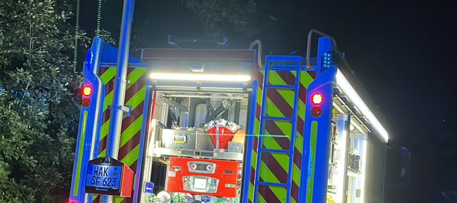 Ein hell erleuchtetes Feuerwehrauto ist nachts auf einer dunklen Straße geparkt. Die hinteren Abteile sind geöffnet und geben den Blick auf die Feuerwehrausrüstung frei. Ein großer Scheinwerfer ist ausgefahren und erhellt die Szene. Im Hintergrund sind Bäume zu sehen.