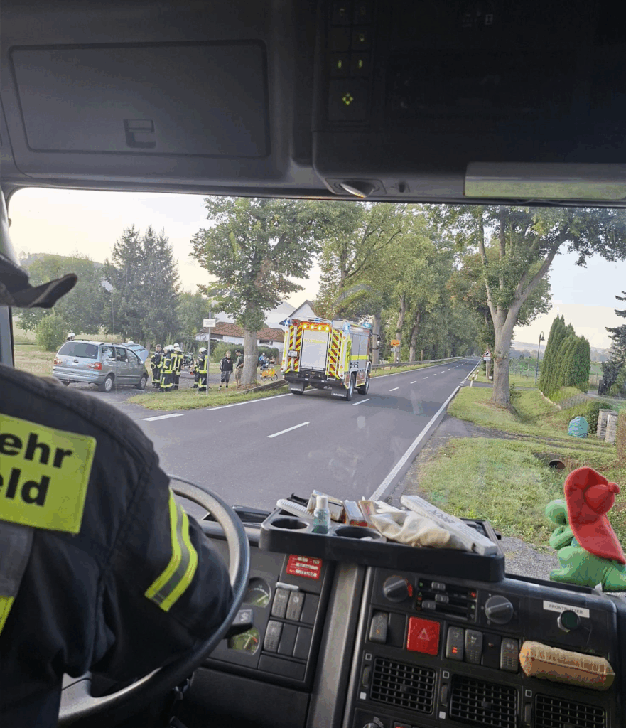 Blick aus dem Inneren eines Feuerwehrautos, das Feuerwehrleute und ein weiteres Feuerwehrauto zeigt, die auf einen Vorfall mit einem Auto am Rande einer Landstraße reagieren. Die Einsatzkräfte sind in der Nähe des Fahrzeugs versammelt, und vor ihnen ist ein Haus zu sehen.