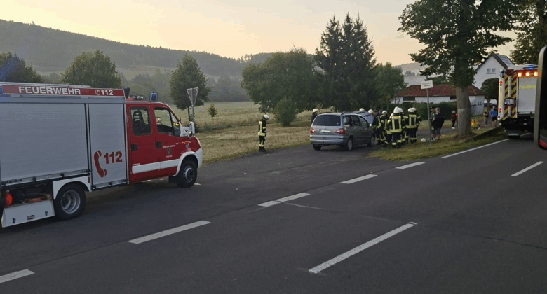 Ein Feuerwehrauto und Rettungskräfte kümmern sich in der Morgendämmerung um einen Autounfall am Rande einer ländlichen Straße. Mehrere Feuerwehrleute stehen in der Nähe eines beschädigten Autos und eines Krankenwagens, umgeben von Bäumen und grasbewachsenen Feldern.