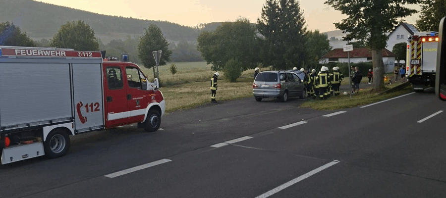 Ein Feuerwehrauto und Rettungskräfte kümmern sich in der Morgendämmerung um einen Autounfall am Rande einer ländlichen Straße. Mehrere Feuerwehrleute stehen in der Nähe eines beschädigten Autos und eines Krankenwagens, umgeben von Bäumen und grasbewachsenen Feldern.