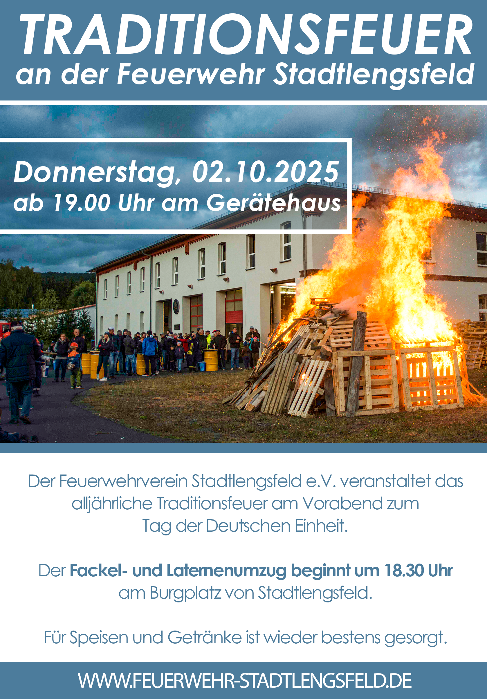 Eine Gruppe von Menschen versammelt sich in der Abenddämmerung vor einer Feuerwache und beobachtet ein großes Lagerfeuer in der Nähe eines Gebäudes. Feuerwehrleute sind anwesend, und ein Banner über ihnen wirbt für ein traditionelles Lagerfeuer zum Tag der Deutschen Einheit.