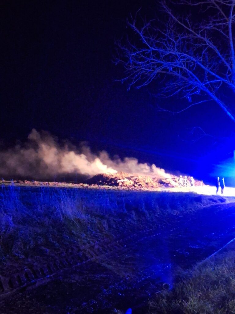 Eine nächtliche Szene zeigt Rauch, der von einem brennenden Haufen Schutt oder Gestrüpp aufsteigt. Blaue Notleuchten erhellen das Gebiet. Zwei Personen stehen in der Nähe des Feuers, und auf der rechten Seite ist ein blattloser Baum zu sehen.