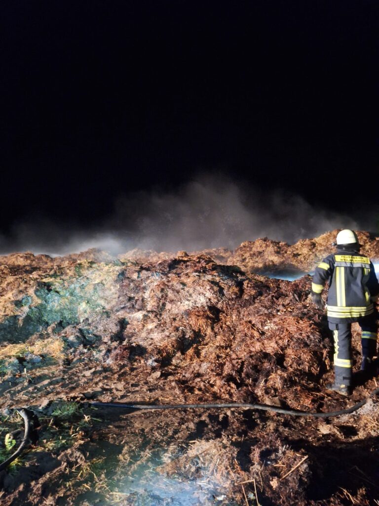 Ein Feuerwehrmann in voller Montur steht nachts in der Nähe eines großen, rauchenden Schutt- oder Komposthaufens, beleuchtet von künstlichem Licht, mit dunklem Himmel im Hintergrund.