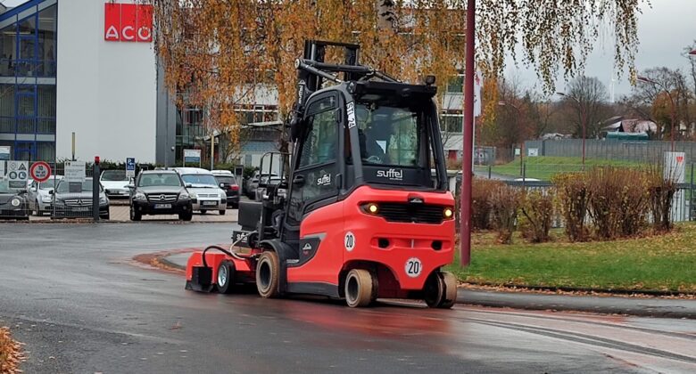 Eine rot-schwarze Straßenreinigungsmaschine fährt auf einer nassen Straße in der Nähe von Herbstbäumen und geparkten Autos, mit Gebäuden im Hintergrund.