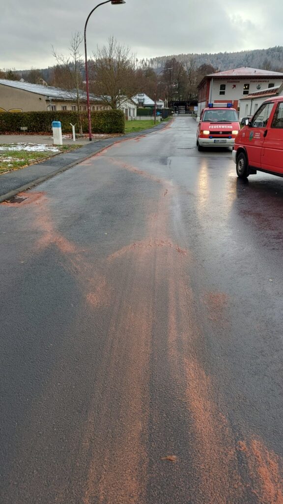 Eine nasse Straße mit rötlich-braunem Pulver, das auf der Oberfläche gestreut wurde, möglicherweise um ausgelaufene Flüssigkeiten aufzufangen. Zwei rote Einsatzfahrzeuge sind auf der rechten Seite geparkt, und auf dem Gras im Hintergrund ist Schnee zu sehen.