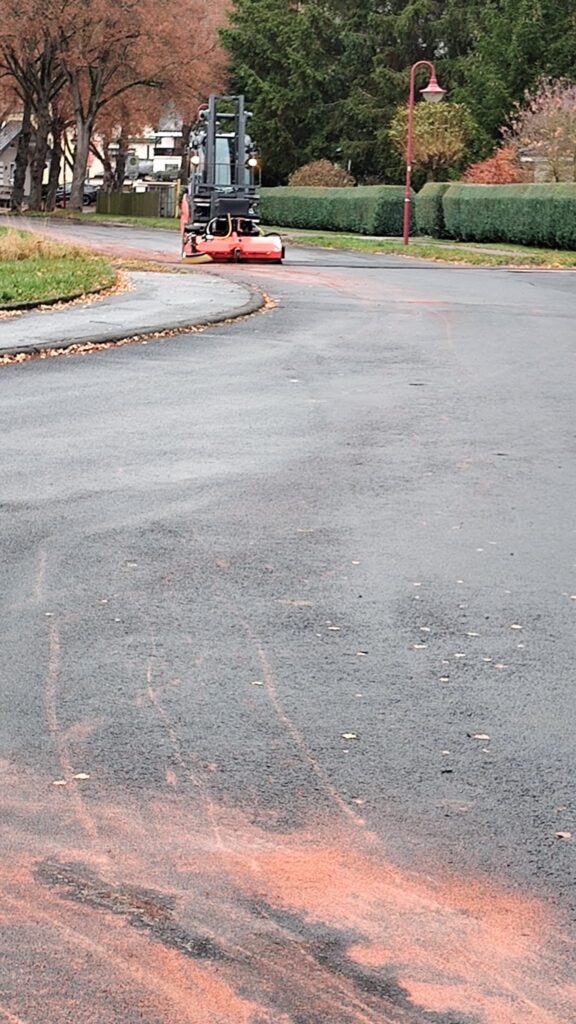Eine Kehrmaschine reinigt eine ruhige, kurvenreiche Straße, die von herbstlichen Bäumen und grünen Hecken gesäumt ist, und hinterlässt eine Spur aus orange-braunem Schmutz auf dem nassen Bürgersteig.