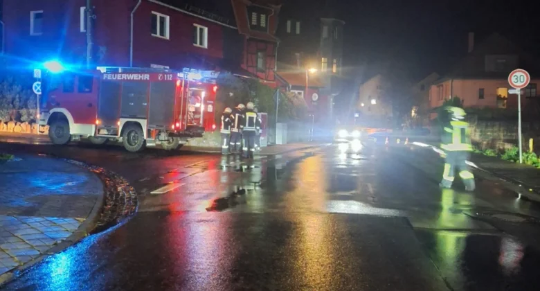 Nachts stehen Feuerwehrleute und ein Löschfahrzeug mit Blinklichtern auf einer nassen Straße vor Häusern. Straßenschilder und Straßenlaternen sind sichtbar, und die nasse Straße reflektiert die Einsatzleuchten.