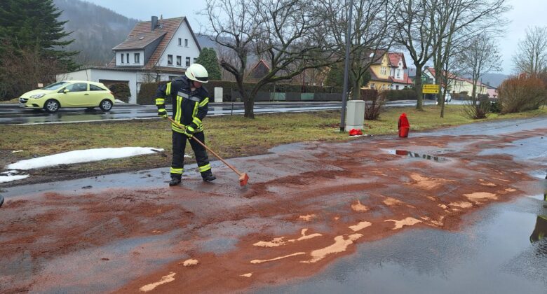 Ein Feuerwehrmann in Uniform fegt an einem bewölkten Tag rötlich-braunes Material von einer nassen Straße in der Nähe eines Bürgersteigs, mit Häusern, Bäumen und einem geparkten gelben Auto im Hintergrund. In der Nähe sind Schneeflecken zu sehen.