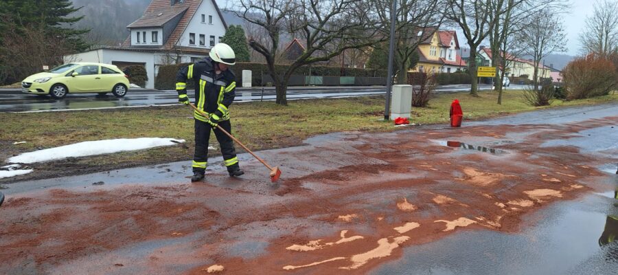 Ein Feuerwehrmann in Uniform fegt an einem bewölkten Tag rötlich-braunes Material von einer nassen Straße in der Nähe eines Bürgersteigs, mit Häusern, Bäumen und einem geparkten gelben Auto im Hintergrund. In der Nähe sind Schneeflecken zu sehen.