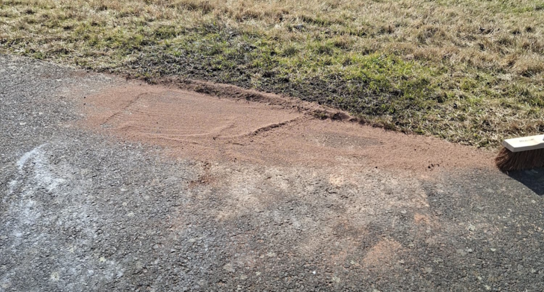 Auf einem asphaltierten Weg neben einer Grasfläche ist ein Stück Sand verteilt. Auf dem Rand des Sandes liegt ein Besen, der andeutet, dass jemand den Sand auf den Weg fegt.