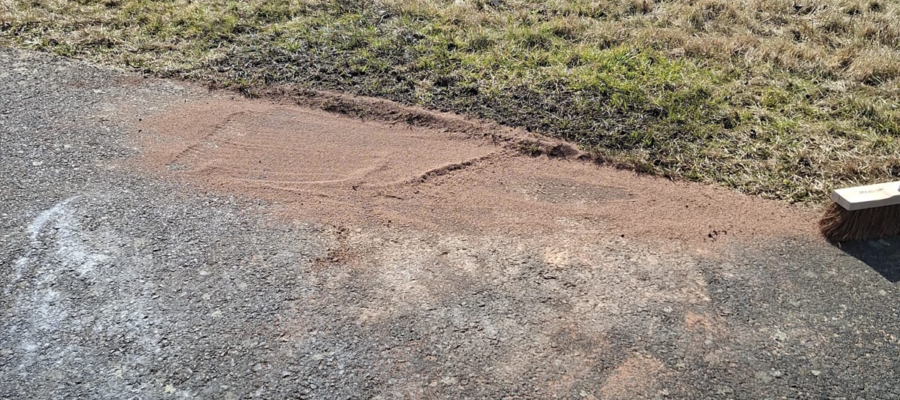 Auf einem asphaltierten Weg neben einer Grasfläche ist ein Stück Sand verteilt. Auf dem Rand des Sandes liegt ein Besen, der andeutet, dass jemand den Sand auf den Weg fegt.