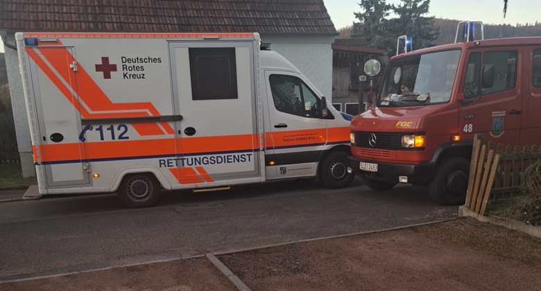 Ein Krankenwagen und ein Feuerwehrauto, beide in deutscher Sprache beschriftet, parken dicht nebeneinander in einer Wohnstraße neben einem Haus und einem Holzzaun.