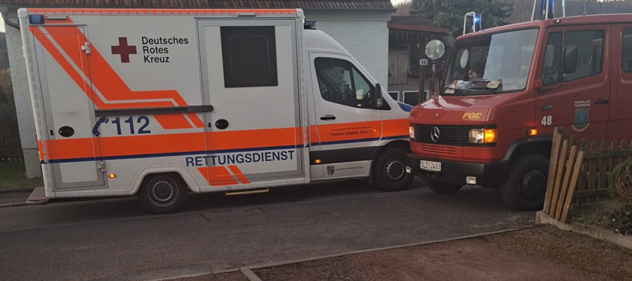 Ein Krankenwagen und ein Feuerwehrauto, beide in deutscher Sprache beschriftet, parken dicht nebeneinander in einer Wohnstraße neben einem Haus und einem Holzzaun.