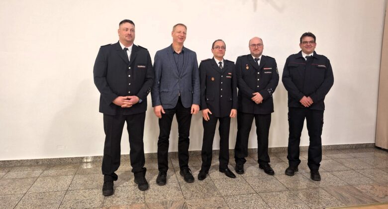 Fünf Männer stehen in einer Reihe in einem Innenraum; vier von ihnen tragen formelle dunkle Uniformen, und einer in der Mitte ist mit einer blauen Jacke, einem schwarzen Hemd und einer schwarzen Hose bekleidet. Sie posieren vor einer schlichten weißen Wand auf einem gefliesten Boden.