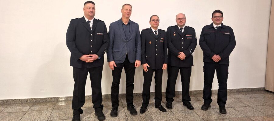 Fünf Männer stehen in einer Reihe in einem Innenraum; vier von ihnen tragen formelle dunkle Uniformen, und einer in der Mitte ist mit einer blauen Jacke, einem schwarzen Hemd und einer schwarzen Hose bekleidet. Sie posieren vor einer schlichten weißen Wand auf einem gefliesten Boden.