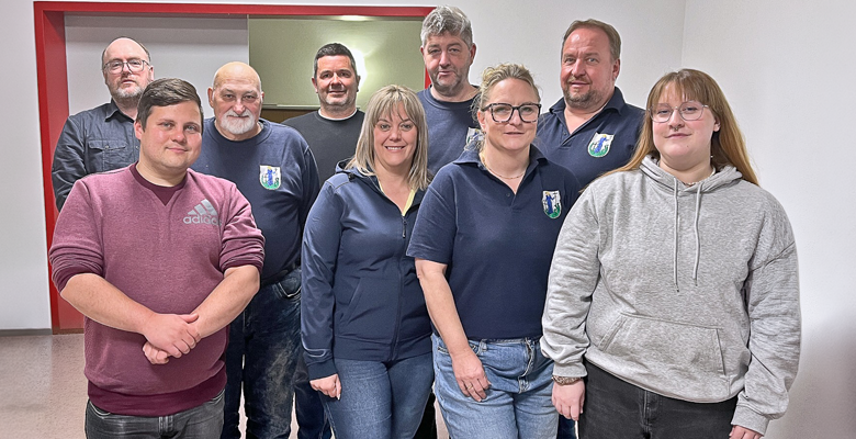 Eine Gruppe von neun Erwachsenen, von denen einige marineblaue Hemden mit einem Wappenlogo tragen, posiert in einem Innenraum vor einer schlichten weißen Wand und einem roten Türrahmen. Die Stimmung ist locker und freundlich.