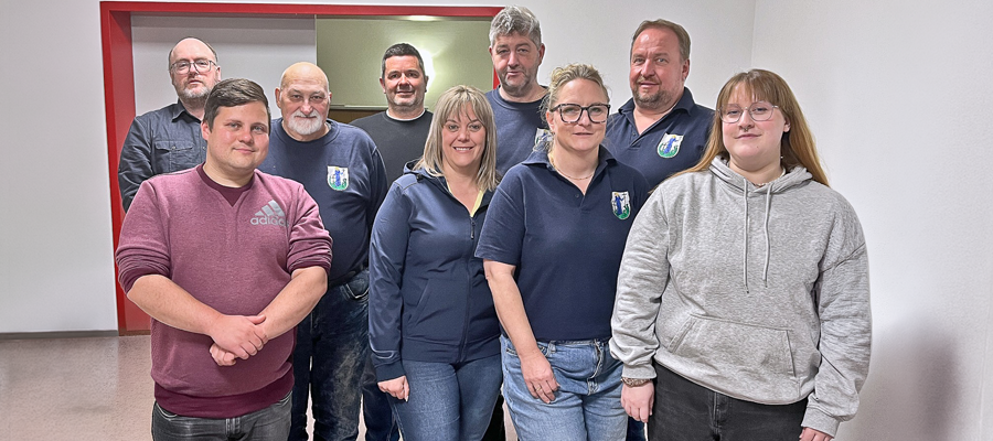 Eine Gruppe von neun Erwachsenen, von denen einige marineblaue Hemden mit einem Wappenlogo tragen, posiert in einem Innenraum vor einer schlichten weißen Wand und einem roten Türrahmen. Die Stimmung ist locker und freundlich.