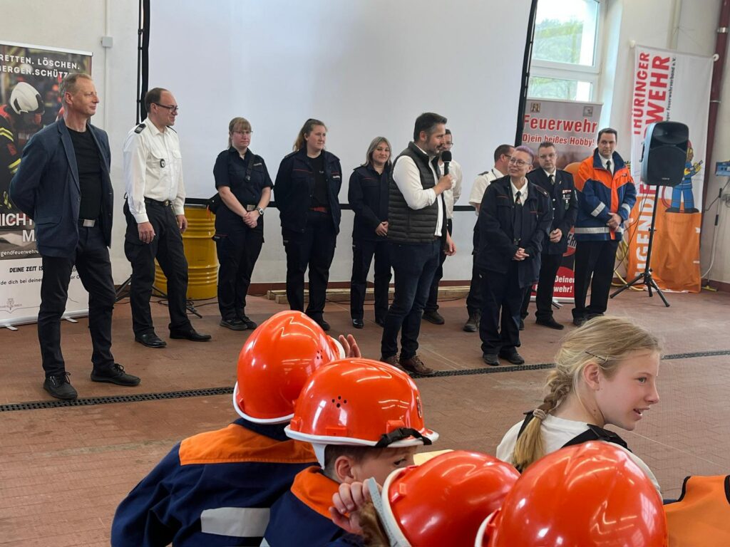 Eine Gruppe von Personen, einige in Feuerwehruniformen und andere in Geschäftskleidung, steht vor einer Präsentationsfläche. Im Vordergrund sitzen Jugendliche in orangefarbenen Helmen und Uniformen und hören aufmerksam zu.