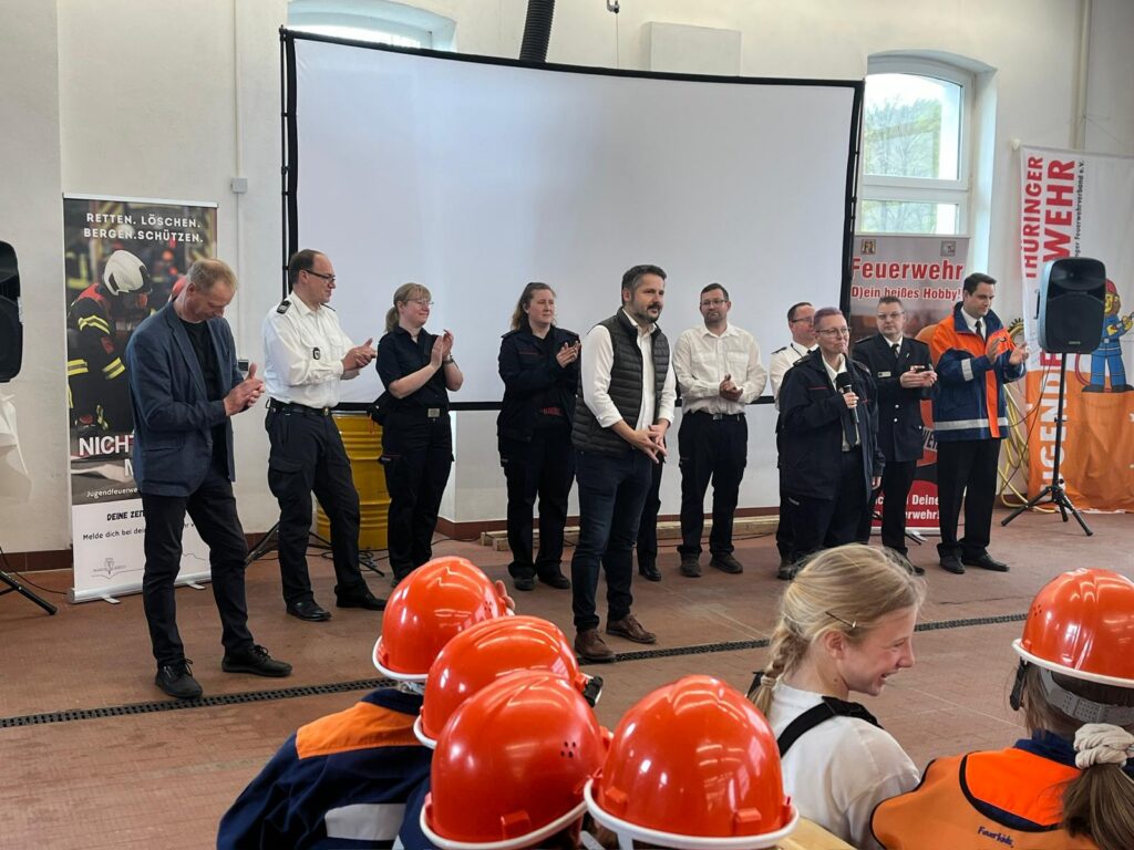 Eine Gruppe Erwachsener steht vor einer Projektionsfläche und applaudiert, während mehrere Kinder in orangefarbenen Feuerwehruniformen und -helmen ihnen gegenüber sitzen. Im Hintergrund sind Feuerwehrfahnen zu sehen.