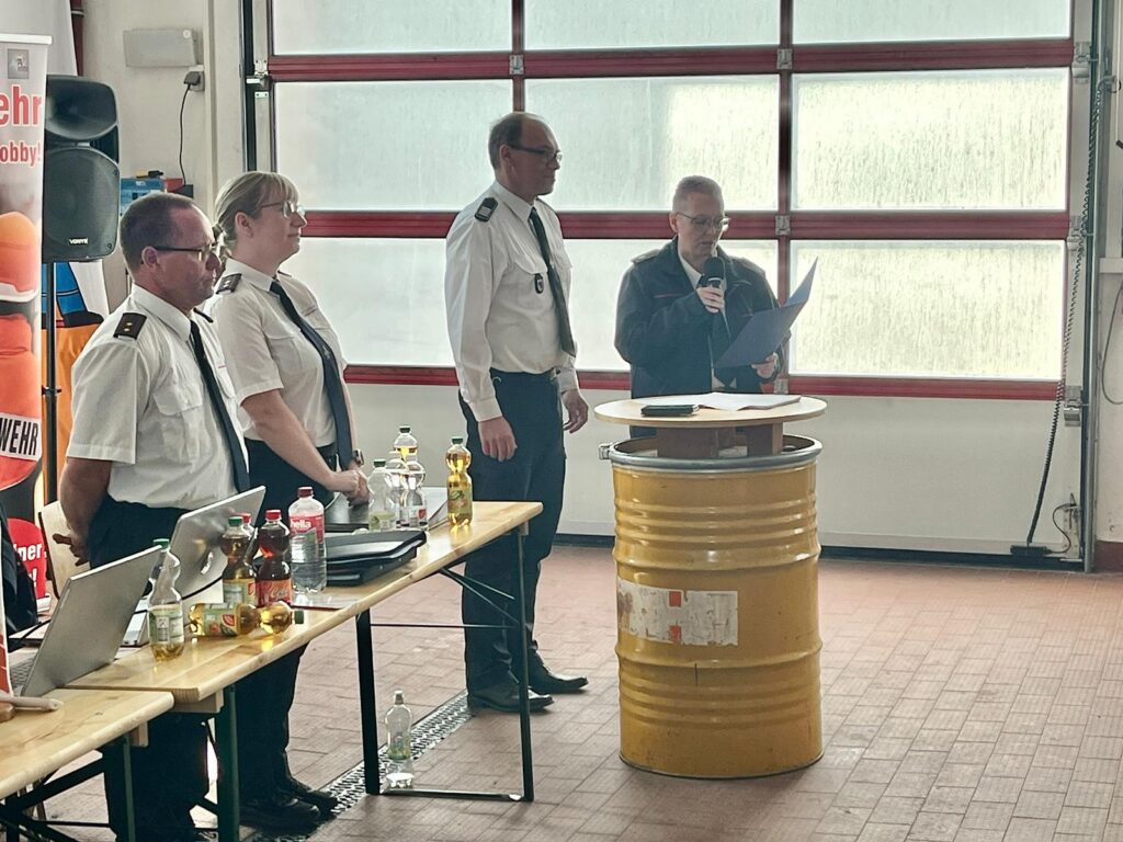 Vier Uniformierte stehen in einem Innenraum in der Nähe einer gelben Tonne, wobei eine Person in ein Mikrofon spricht und Papiere in der Hand hält. Neben ihnen ist ein Tisch mit Getränken und einem Laptop vor großen, garagenartigen Fenstern zu sehen.