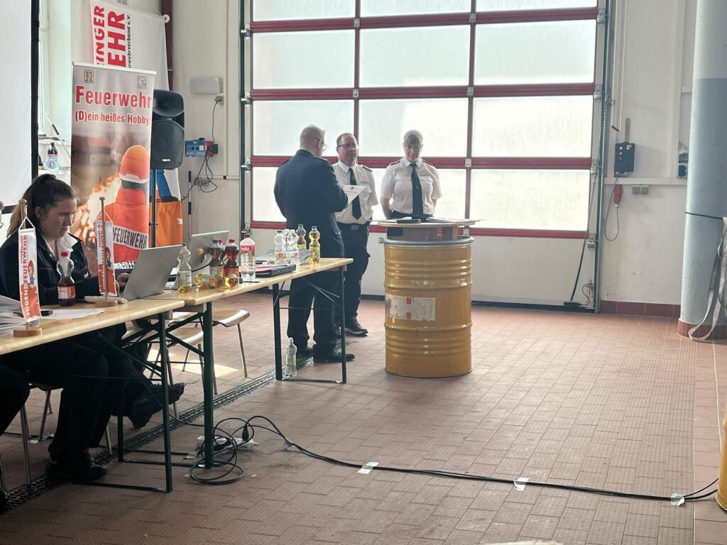 Drei Uniformierte stehen und sprechen neben einem gelben Podium in einer Feuerwache. Auf der linken Seite sitzen andere an einem Tisch mit Laptops, Trophäen und Bannern zum Thema Brandschutz im Hintergrund.