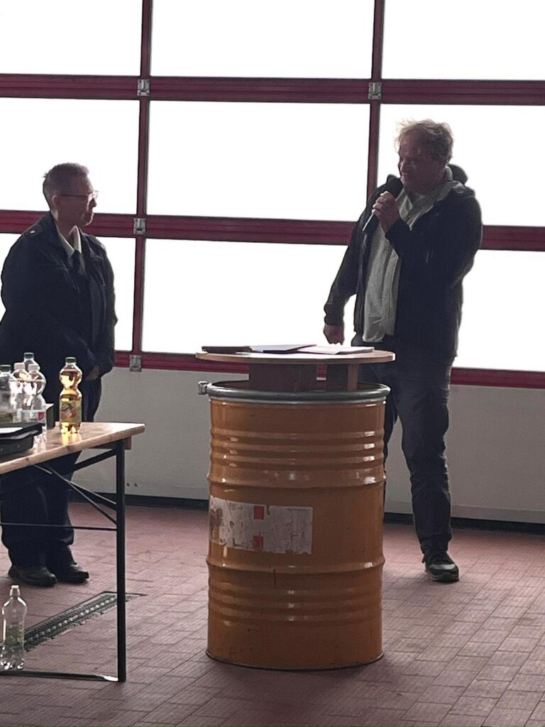 Zwei Personen stehen in einem Raum neben einem gelben Fass, das als Podium dient. Die eine spricht in ein Mikrofon, während die andere zuhört. In der Nähe befinden sich Flaschen und ein Tisch mit Gegenständen, und im Hintergrund ist ein großes Fenster mit rotem Rahmen zu sehen.