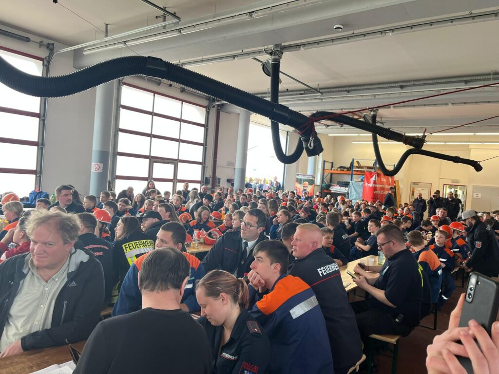 Eine große Gruppe von Menschen, meist in orangefarbenen und blauen Uniformen, sitzt an Tischen in einem geräumigen, gut beleuchteten Innenraum und scheint an einer Veranstaltung oder Sitzung teilzunehmen. Auf einigen Uniformen ist Feuerwehr aufgedruckt.