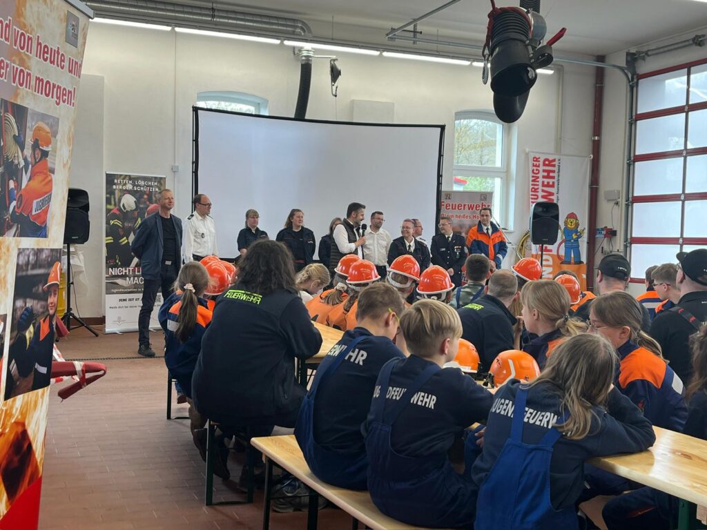 Eine Gruppe von Menschen steht und spricht vor einer Projektionsfläche, während Kinder und Jugendliche in Feuerwehruniformen und -helmen an Tischen in einem hellen Innenraum sitzen.