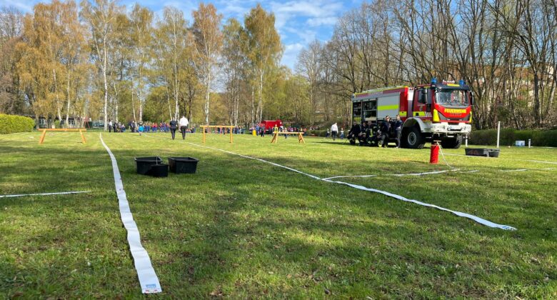 Ein grasbewachsenes Feld mit auf dem Boden ausgelegten Feuerwehrschläuchen, einem Feuerwehrauto und Menschen in Uniformen. Im Hintergrund sind Bäume mit Frühlingsblättern und eine Gruppe von Menschen unter einem blauen Himmel mit dünnen Wolken zu sehen.