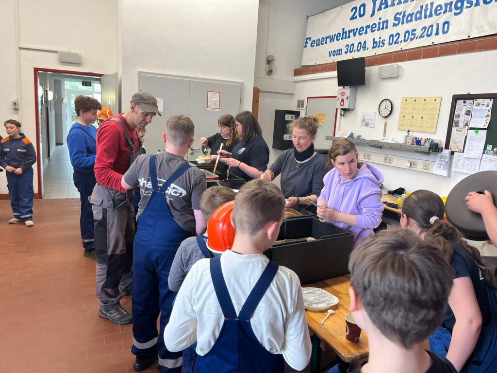 Eine Gruppe von Kindern und Erwachsenen versammelt sich in einem Raum, einige tragen Overalls und Arbeitsuniformen und bereiten gemeinsam an Tischen das Essen zu. Es gibt Küchenutensilien und ein Schild an der Wand mit deutschem Text und Daten.