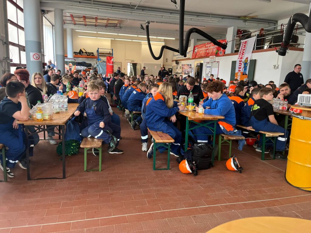 Eine große Gruppe von Menschen, meist Kinder und Jugendliche in blauen Uniformen, sitzt an langen Holztischen in einer hellen Halle, isst und unterhält sich. Schutzhelme liegen auf dem Boden oder auf den Tischen. Die Atmosphäre ist lebhaft.