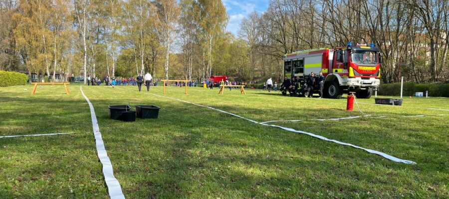 Ein grasbewachsenes Feld mit auf dem Boden ausgelegten Feuerwehrschläuchen, einem Feuerwehrauto und Menschen in Uniformen. Im Hintergrund sind Bäume mit Frühlingsblättern und eine Gruppe von Menschen unter einem blauen Himmel mit dünnen Wolken zu sehen.