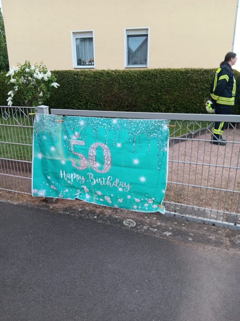 Ein blaugrünes Transparent mit silbernen Verzierungen und dem Text 50 Happy Birthday hängt an einem Metallzaun vor einem Haus. Eine Person in einer Feuerwehruniform steht im Hintergrund in der Nähe einer Hecke.