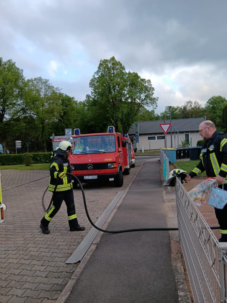 Zwei Feuerwehrmänner in Ausrüstung stehen in der Nähe eines Bürgersteigs; einer hält einen Schlauch, während der andere an einem Zaun steht. Ein rotes Feuerwehrauto ist an einem bewölkten Tag in der Nähe geparkt, mit Bäumen und Gebäuden im Hintergrund.