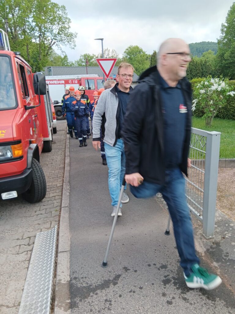 Eine Gruppe von Menschen, darunter Feuerwehrleute in Uniform, geht neben roten Feuerwehrautos einen Bürgersteig entlang. Ein lächelnder Mann mit Krücken geht voran, gefolgt von anderen, mit Bäumen und Grünflächen im Hintergrund.