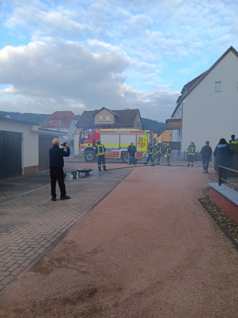 Feuerwehrleute in voller Montur stehen neben einem Löschfahrzeug in einer Wohnstraße, während eine Person die Szene fotografiert. Im Hintergrund ist Rauch zu sehen, und mehrere Personen beobachten die Szene aus der Ferne in der Nähe von Häusern.