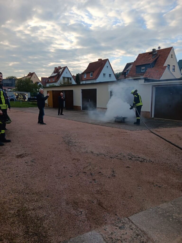 Ein Feuerwehrmann in voller Montur sprüht Wasser auf ein rauchendes Objekt vor einer Garage, während drei Personen das Geschehen beobachten, mit Häusern und einem bewölkten Himmel im Hintergrund.