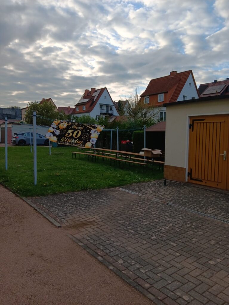 Ein Hinterhof mit einem langen Tisch und Bänken neben einem Transparent mit der Aufschrift 50. Geburtstag, geschmückt mit goldenen und weißen Luftballons; rote Häuser mit Dächern und ein bewölkter Himmel im Hintergrund.