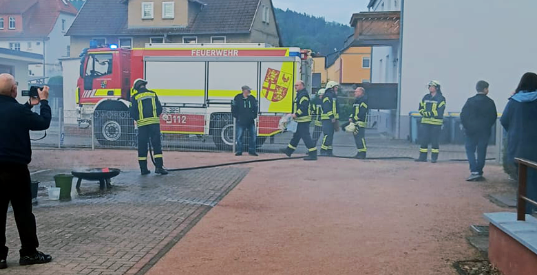 Feuerwehrleute in gelben Anzügen stehen neben einem Feuerwehrauto mit der Aufschrift "Feuerwehr". Rauch liegt in der Luft, während eine Person ein Foto macht, und andere die Szene in einem Wohngebiet beobachten.