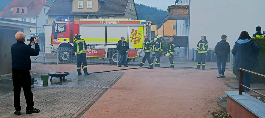 Feuerwehrleute in gelben Anzügen stehen neben einem Feuerwehrauto mit der Aufschrift "Feuerwehr". Rauch liegt in der Luft, während eine Person ein Foto macht, und andere die Szene in einem Wohngebiet beobachten.