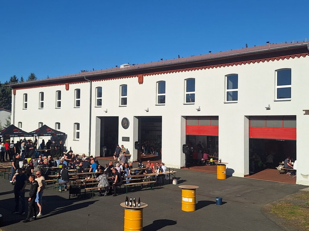 Vor einem weißen Gebäude mit großen Garagentoren sind Menschen versammelt, die an Picknicktischen sitzen und den sonnigen Tag genießen. Links im Bild stehen gelbe Fässer, die als Tische dienen, und ein Zelt.