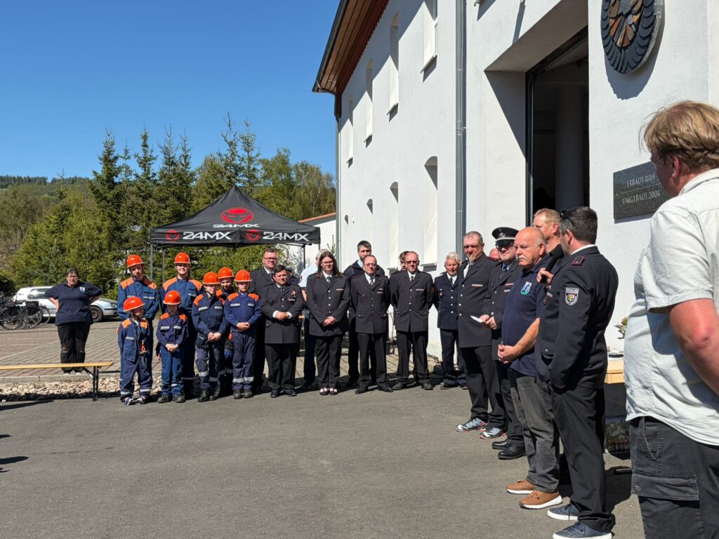 Eine Gruppe von Menschen, darunter Feuerwehrleute und Beamte in Uniform, steht an einem sonnigen Tag in einer Reihe vor einem weißen Gebäude. Einige tragen blaue Uniformen mit orangefarbenen Helmen, während andere dunkle Anzüge tragen. Im Hintergrund sind ein Zelt und Bäume zu sehen.