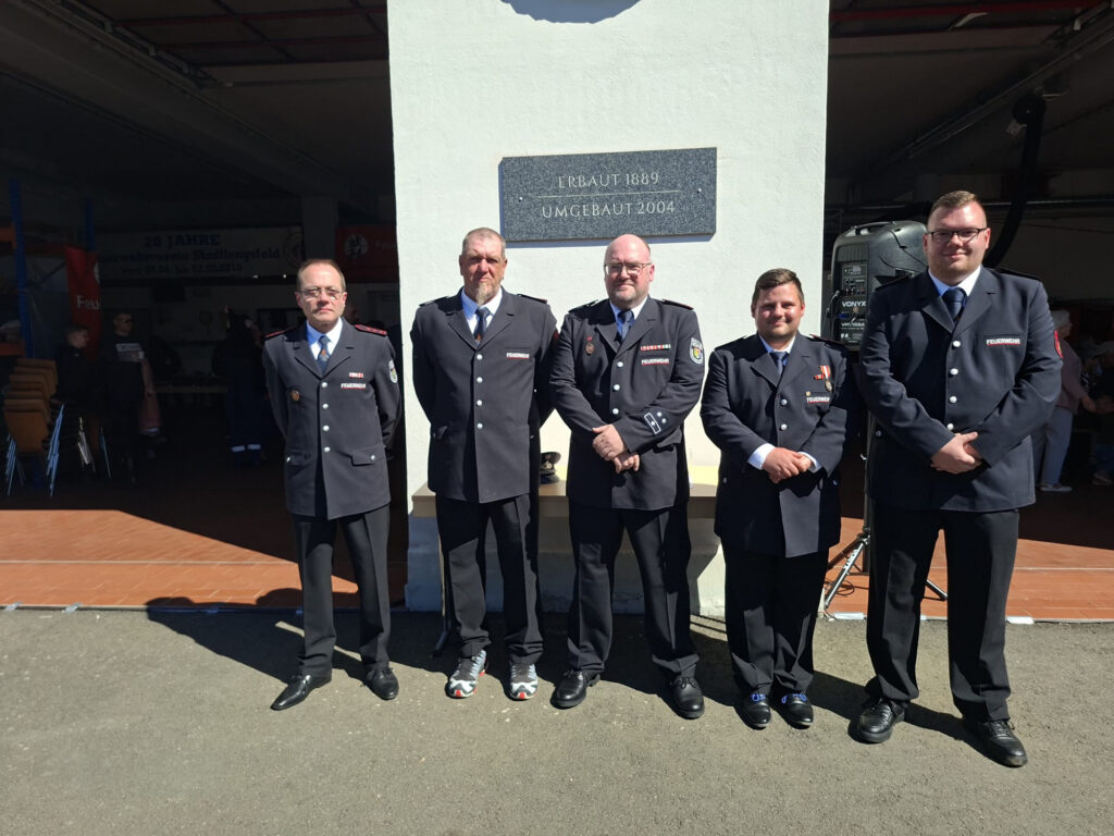 Fünf Männer in Uniform stehen in einer Reihe vor einem Gebäude und schauen in die Kamera. Sie stehen im Sonnenlicht, und an der Wand hinter ihnen ist ein Schild angebracht. Im Inneren des Gebäudes sind Tische und Stühle zu sehen.