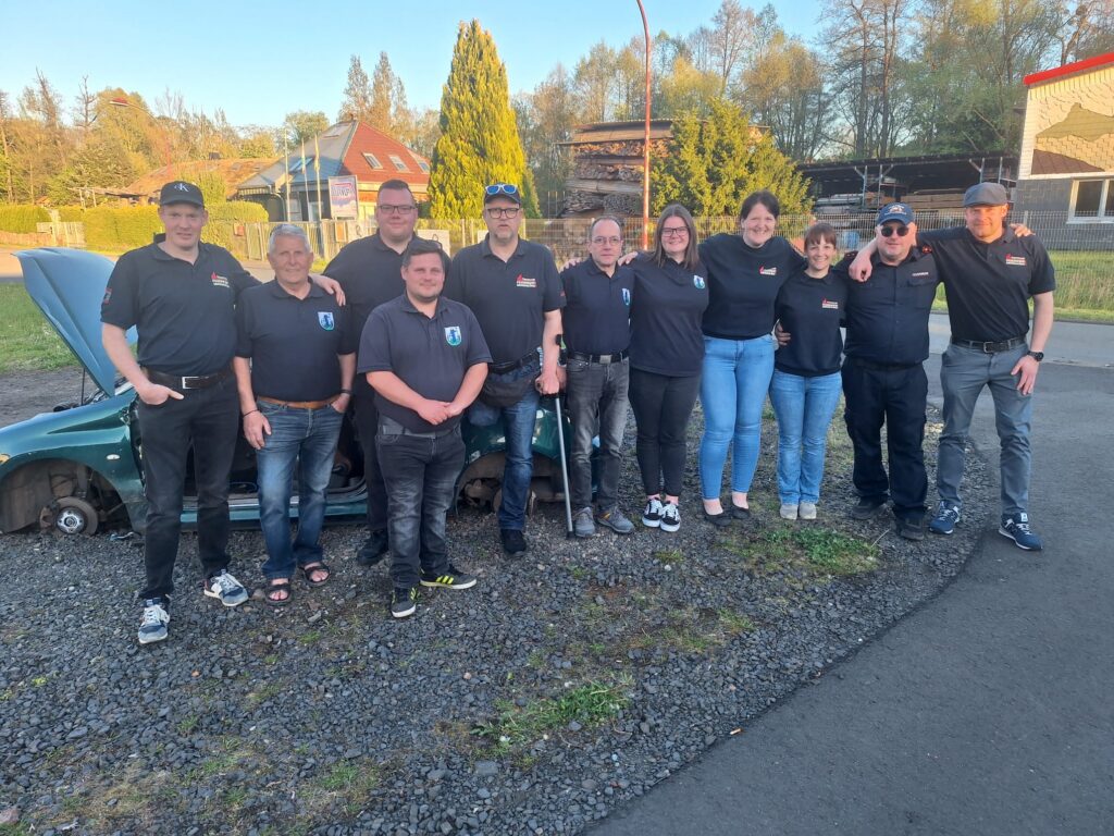 Eine Gruppe von zehn Personen, meist in passenden schwarzen Hemden und Jeans, steht an einem sonnigen Tag im Freien zusammen und lächelt, während im Hintergrund Grünflächen, ein teilweise sichtbares Auto und Gebäude zu sehen sind.