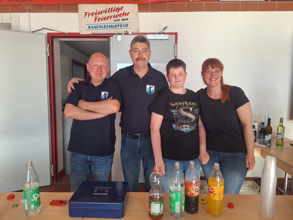 Vier Personen stehen hinter einem Tresen mit Softdrinks und Flaschen. Drei tragen zusammenpassende marineblaue Hemden mit einem Wappen, und ein junger Mensch trägt ein schwarzes T-Shirt mit Grafik. Sie stehen lächelnd in einem hell erleuchteten Raum mit einem Schild über ihnen.